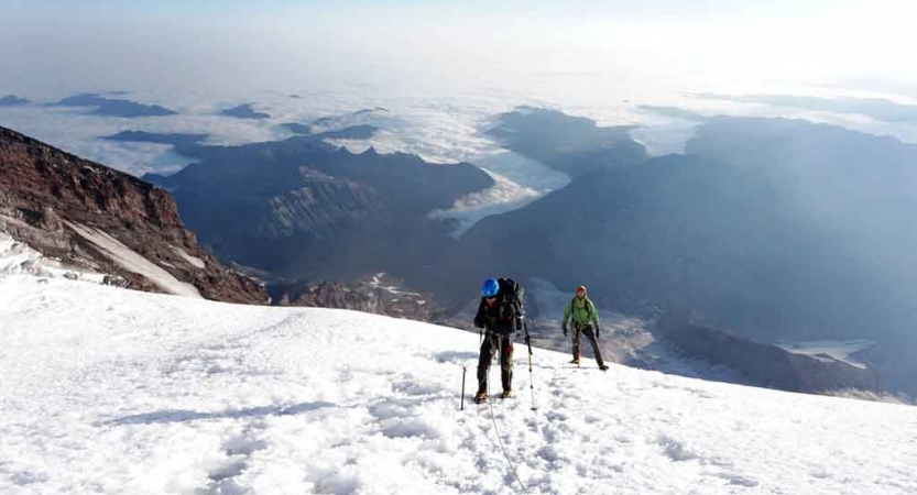 two people wearing mountaineering gear stand on a very high snowy field overlooking a vast mountainous landscape